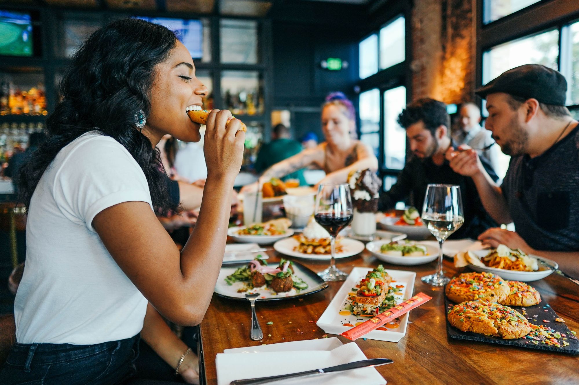 Mulher preta de blusa branca comendo um pedaco de pao numa mesa com um homem obeso, um homem magro e uma mulher branca. Tod