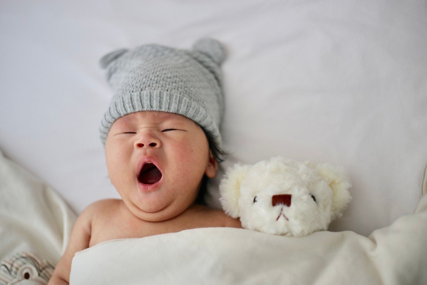 Bebê com gorro cinza boceja e estádeitado em lençol bege de olhos fechados ao lado de ursinho creme.