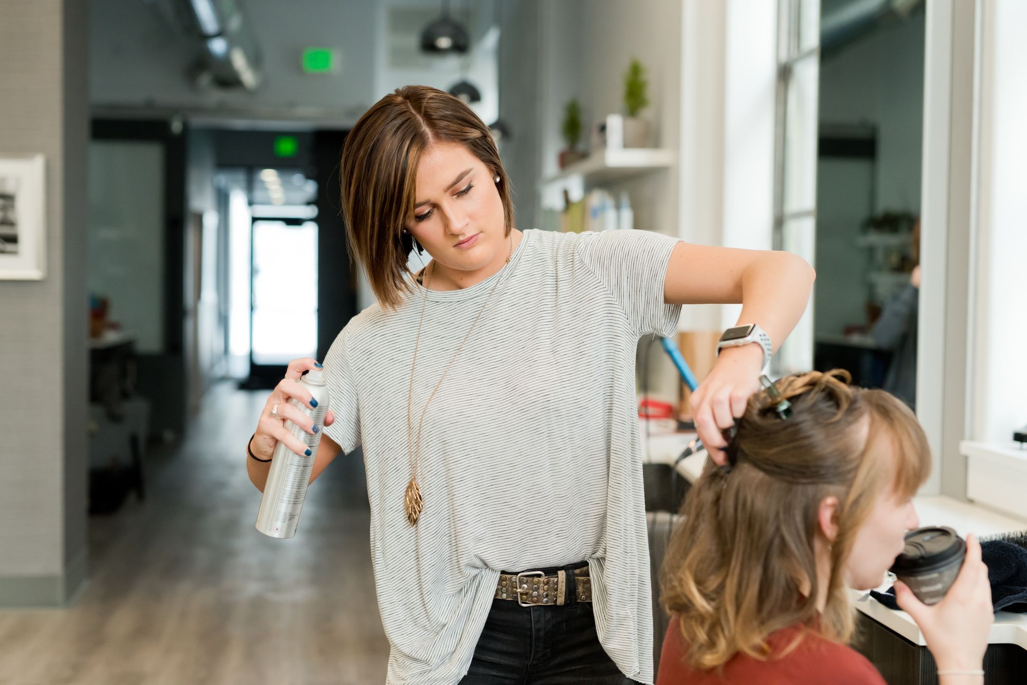 Mulher em pé faz penteado em cabelo de outra mulher que toma café sentada.